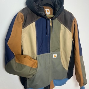 Pode incluir: Um casaco com capuz em blocos de cor com painéis em castanho, azul-marinho, verde-azeitona, bege e azul-claro. O casaco tem um capuz preto acolchoado, fecho de correr frontal e um emblema da marca Carhartt no peito e no interior da gola.