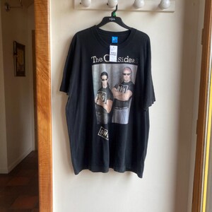 May include: Black t-shirt featuring a graphic of two people wearing sunglasses with the text "The Outsiders" above them. The shirt has a tag attached and is hanging on a black hanger.