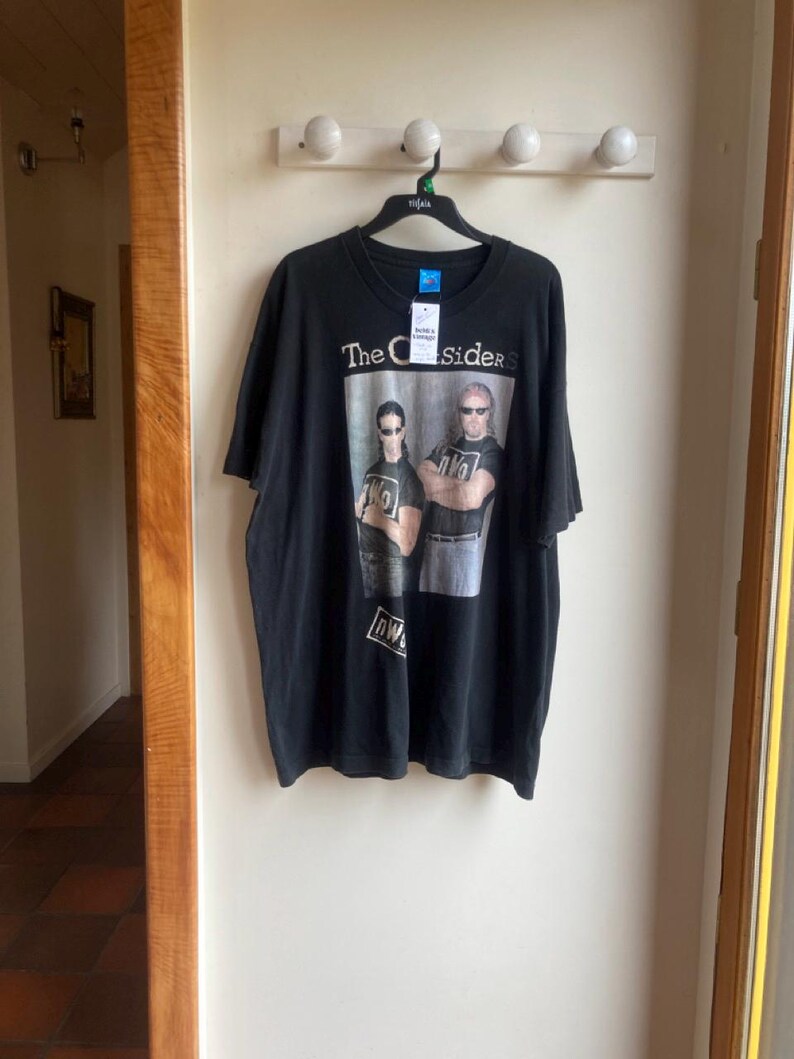 May include: Black t-shirt featuring a graphic of two people wearing sunglasses with the text "The Outsiders" above them. The shirt has a tag attached and is hanging on a black hanger.
