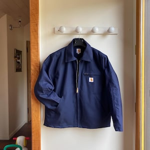 Könnte beinhalten: Marineblaue Arbeitsjacke mit durchgehendem Reißverschluss und Kragen. Die Jacke hat zwei Vordertaschen und einen kleinen quadratischen Logoaufnäher auf der Brust. Sie hängt an einer weißen Wand.