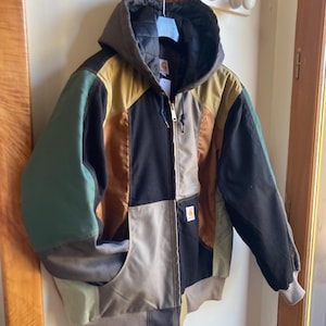 Peut inclure: Veste à capuche patchwork aux tons de vert, marron, noir et beige. La veste est dotée d'une fermeture éclair intégrale, d'une capuche à cordon de serrage et de deux poches avant. Le logo Carhartt est visible.