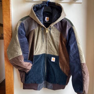 Op de afbeelding: Een veelkleurige corduroy jas met capuchon. De jas heeft een patchwork ontwerp in tinten bruin, beige, marineblauw en olijfgroen. Het heeft een ritssluiting en een klein logopatch op de voorkant. De jas hangt aan een muur.