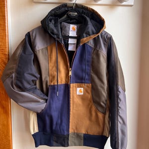 Könnte beinhalten: Eine farblich abgesetzte Kapuzenjacke mit Einsätzen in Braun, Marineblau, Anthrazit und Beige. Die Jacke verfügt über einen Frontreißverschluss, eine Kapuze mit Kordelzug, gerippte Bündchen und ein Carhartt-Markenlogo auf der linken Brust.
