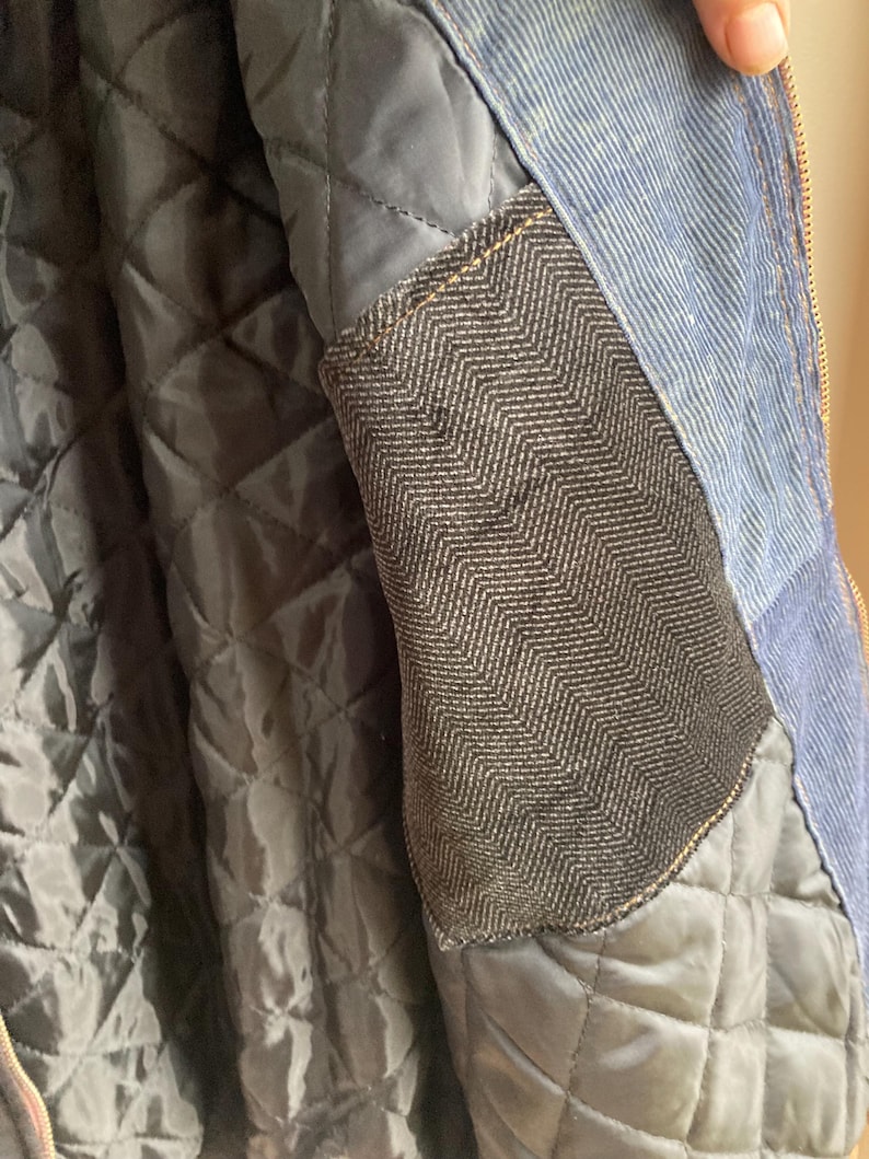 May include: Close-up of a blue denim jacket with a quilted black lining. The jacket features a dark gray herringbone pocket and a gold zipper. The interior lining is a diamond pattern.