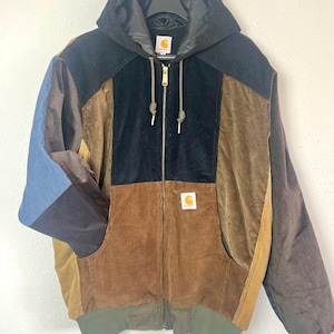 Pode incluir: Um casaco com capuz em veludo cotelê com blocos de cores, apresentando um fecho de correr frontal completo, punhos e bainha canelados e um emblema da marca Carhartt no peito. A peça é construída com painéis de tecido de veludo cotelê preto, castanho, bege e azul.
