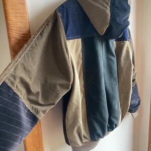 May include: A multi-colored corduroy jacket with a hood. The jacket features panels of brown, navy blue, and black corduroy. The sleeves have a striped pattern. The jacket is hanging on a wall.