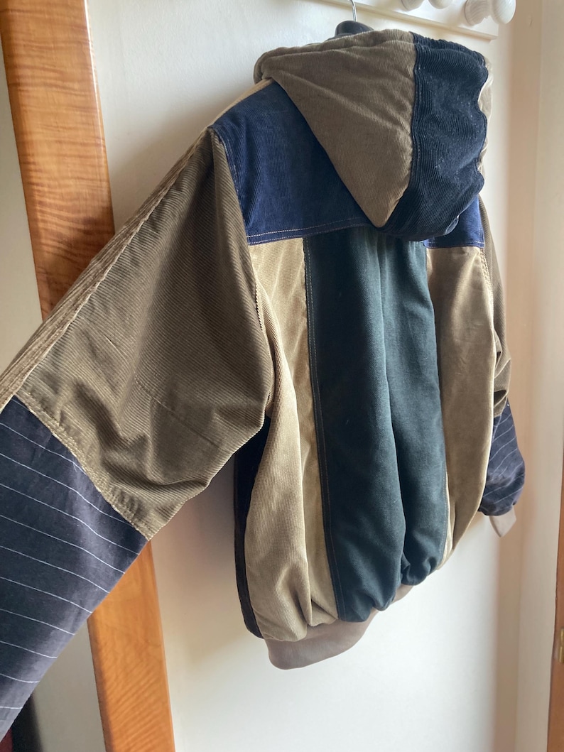 May include: A multi-colored corduroy jacket with a hood. The jacket features panels of brown, navy blue, and black corduroy. The sleeves have a striped pattern. The jacket is hanging on a wall.