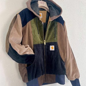 Op de afbeelding: Een color-block corduroy jack met capuchon, voorzien van panelen in bruin, olijfgroen, marineblauw en beige. Het jack heeft een ritssluiting aan de voorzijde, een capuchon met trekkoord, geribbelde boorden en een Carhartt-logo op de borst.