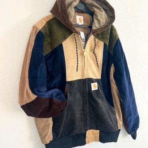 Könnte beinhalten: Eine farblich abgesetzte Kapuzenjacke aus Cord mit durchgehendem Frontreißverschluss, langen Ärmeln und gerippten Bündchen. Das Kleidungsstück besteht aus Stoffbahnen in Braun, Marineblau, Beige, Olivgrün und Schwarz mit einem Carhartt-Markenetikett.