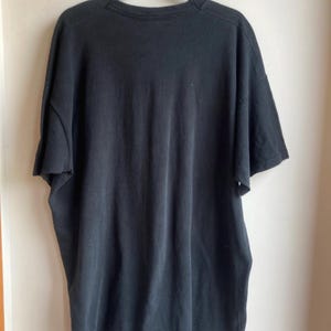 May include: A black short-sleeved t-shirt hanging on a black hanger. The shirt is made of a soft, dark fabric and appears to be slightly worn. The t-shirt is hanging against a white wall.