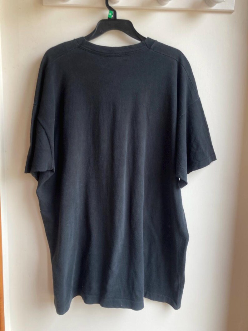 May include: A black short-sleeved t-shirt hanging on a black hanger. The shirt is made of a soft, dark fabric and appears to be slightly worn. The t-shirt is hanging against a white wall.