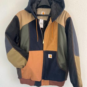 May include: A colour-blocked hooded jacket featuring panels of tan, olive green, and navy blue fabric. The jacket has a front zip closure, a quilted hood lining, ribbed cuffs and hem, and a Carhartt brand patch on the chest and inner neck.