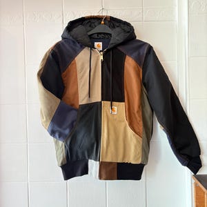 Op de afbeelding: Een patchwork jack met capuchon in diverse kleuren, waaronder zwart, bruin, beige en marineblauw. De jas heeft een ritssluiting aan de voorkant, een capuchon en een Carhartt-logopatch op de zak.