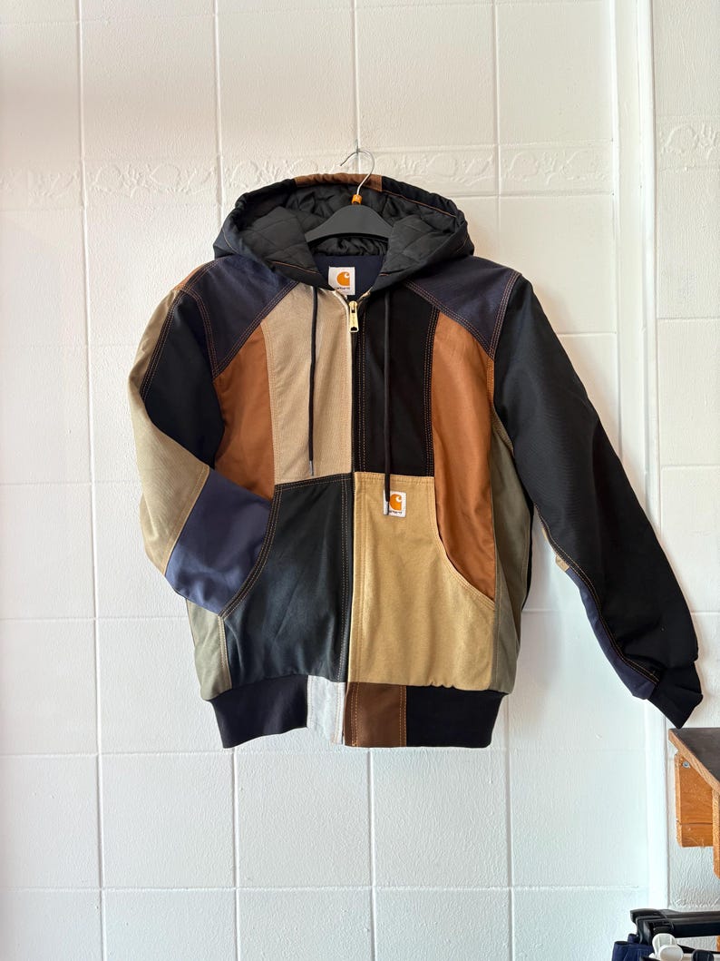 May include: A patchwork hooded jacket with a variety of colors including black, brown, tan, and navy blue. The jacket has a front zipper, a hood, and a Carhartt logo patch on the pocket.