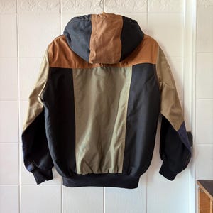 May include: A hooded jacket with a color block design. The jacket features a combination of tan, brown, olive green, and black panels. The jacket is hanging on a metal hanger against a white tiled wall.