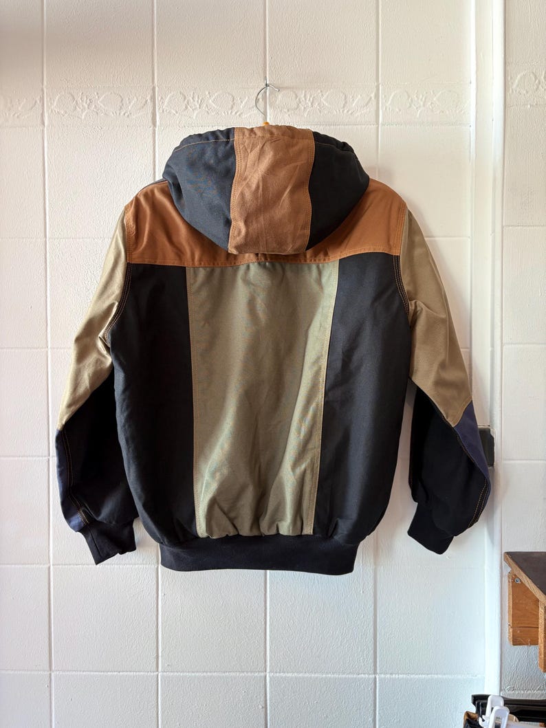 May include: A hooded jacket with a color block design. The jacket features a combination of tan, brown, olive green, and black panels. The jacket is hanging on a metal hanger against a white tiled wall.