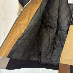 May include: A tan, brown, and black bomber jacket with a quilted black lining. The jacket features a zipper closure and a contrasting black hem and cuffs. The jacket has a vintage aesthetic.