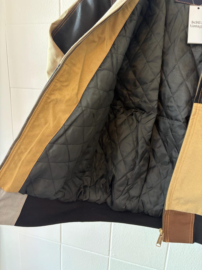 May include: A tan, brown, and black bomber jacket with a quilted black lining. The jacket features a zipper closure and a contrasting black hem and cuffs. The jacket has a vintage aesthetic.