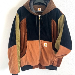 Könnte beinhalten: Ein farblich abgesetzter Cord-Kapuzenpullover mit Reißverschluss, brauner Kapuze, schwarzen oberen Einsätzen und rostorangen unteren Einsätzen sowie olivgrünen Ärmelakzenten. Die Jacke verfügt über einen Frontreißverschluss und ein Carhartt-Label.