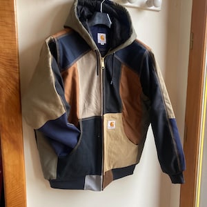 May include: A patchwork design hooded jacket with a zip closure and two front pockets. The jacket features a Carhartt logo patch. The colour scheme includes black, brown, tan, and navy blue. The jacket is hanging on a wall.