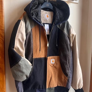 May include: A colour-blocked hooded jacket featuring panels of black, tan, olive green, and brown fabric. The jacket has a front zip closure, a quilted black hood lining, ribbed cuffs, and a Carhartt brand patch on the chest and inner neck label.