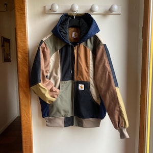 Może przedstawiać: Kurtka patchwork z kapturem, w różnych kolorach, w tym granatowym, brązowym, oliwkowym i beżowym. Kurtka ma zapięcie na zamek błyskawiczny i małą naszywkę z logo. Wisi na białej ścianie.