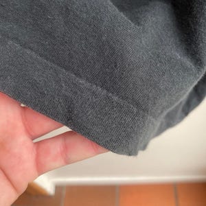 May include: Close-up of a dark gray fabric, possibly a t-shirt or other garment. The fabric is folded, revealing the hem and texture of the material. The image is well-lit, highlighting the details of the textile.