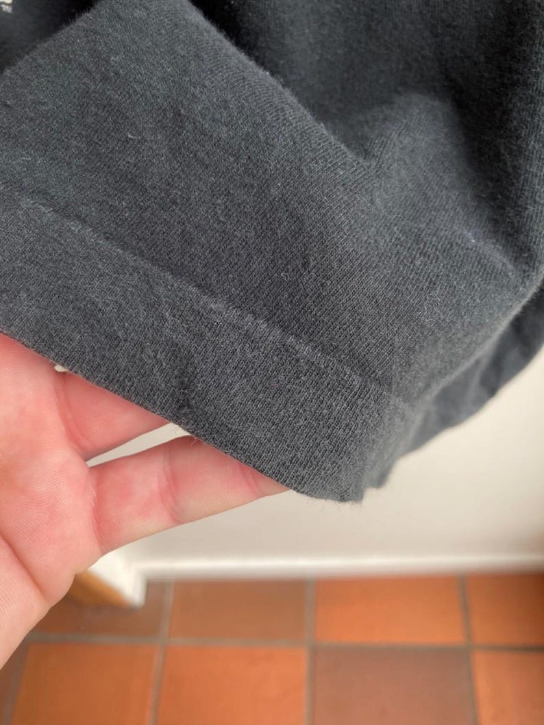 May include: Close-up of a dark gray fabric, possibly a t-shirt or other garment. The fabric is folded, revealing the hem and texture of the material. The image is well-lit, highlighting the details of the textile.