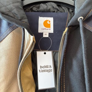 May include: A Carhartt jacket with a black quilted lining and a beige, navy, and black exterior. The jacket has a zipper closure and a tag that reads "beldi's Vintage". The Carhartt logo is visible on the label.