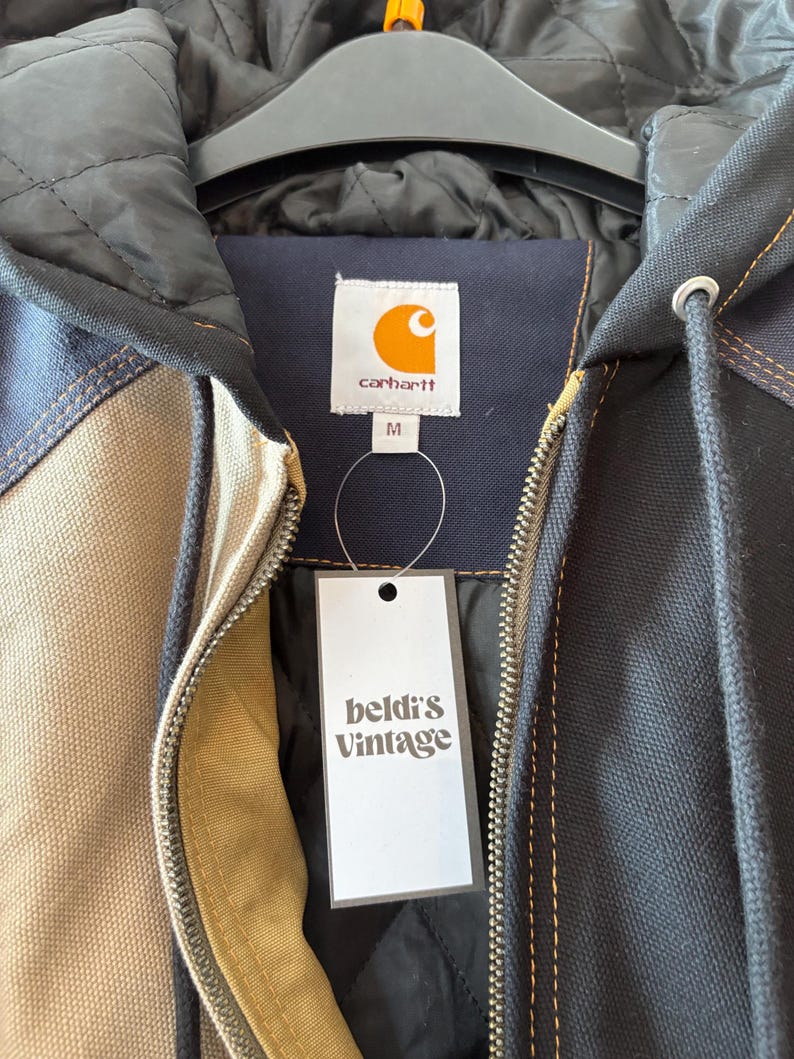 May include: A Carhartt jacket with a black quilted lining and a beige, navy, and black exterior. The jacket has a zipper closure and a tag that reads "beldi's Vintage". The Carhartt logo is visible on the label.