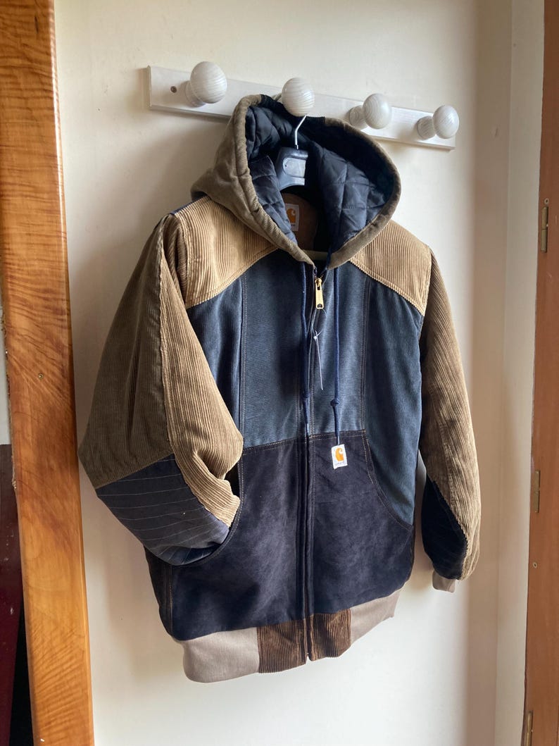 May include: A multi-toned corduroy jacket with a hood, hanging on a white wall. The jacket features panels of brown, blue, and black corduroy. It has a full zipper closure and a small logo tag. The jacket is approximately 28 inches long.