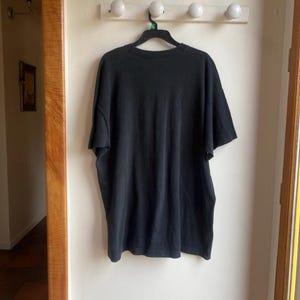 May include: A black short-sleeved t-shirt hanging on a black hanger. The shirt is made of a soft material and is hanging on a white wall. A wooden door frame is visible on the left and right sides of the image.