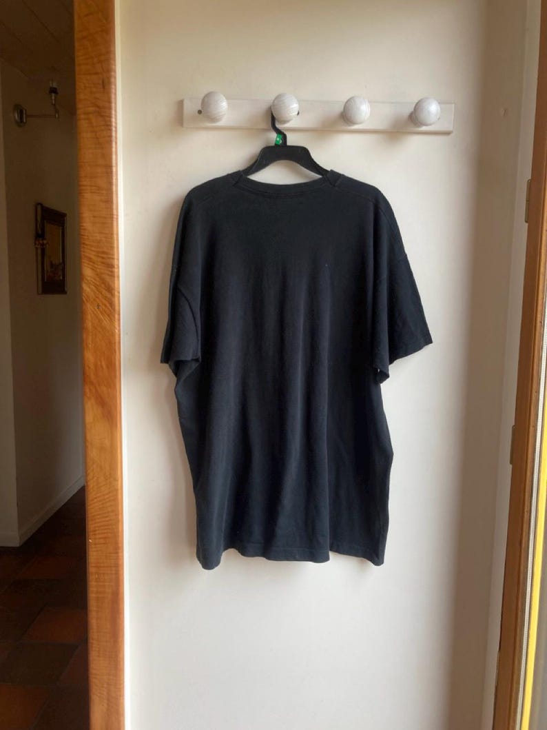 May include: A black short-sleeved t-shirt hanging on a black hanger. The shirt is made of a soft material and is hanging on a white wall. A wooden door frame is visible on the left and right sides of the image.