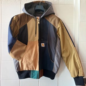 Op de afbeelding: Een veelkleurige jas met capuchon en ritssluiting. De jas heeft panelen in bruin, beige, zwart, grijs en marineblauw. Het heeft een zwarte geribbelde zoom en manchetten en een kleine logopatch aan de voorkant.