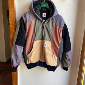 Op de afbeelding: Een veelkleurige Carhartt jas met capuchon en ritssluiting. De jas heeft panelen van zwarte, paarse, bruine en roestkleurige stof. Het heeft een capuchon, twee voorzakken en een Carhartt-logopatch.