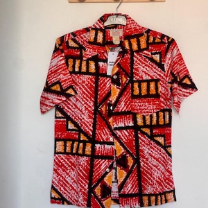 Può includere: Una camicia a maniche corte con bottoni caratterizzata da un audace motivo geometrico in rosso, bianco, nero e arancione. La camicia presenta un colletto a punta, chiusura frontale con bottoni e una tasca sul petto. Un'etichetta bianca è attaccata al colletto.