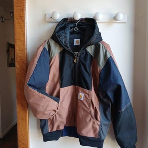 Può includere: Giacca Carhartt multicolore con cappuccio, appesa a una parete bianca. La giacca presenta un design a blocchi di colore con pannelli marroni, blu navy, neri e verde oliva. Ha una chiusura con cerniera completa e due tasche anteriori. Il logo Carhartt è visibile sul petto.