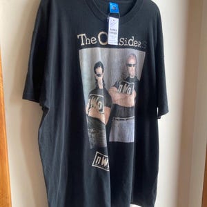 May include: Black t-shirt featuring the text "The Outsiders" above a graphic of two men wearing sunglasses. The graphic also includes the letters "nWo". The shirt is hanging on a black hanger.
