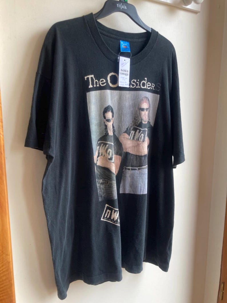 May include: Black t-shirt featuring the text "The Outsiders" above a graphic of two men wearing sunglasses. The graphic also includes the letters "nWo". The shirt is hanging on a black hanger.