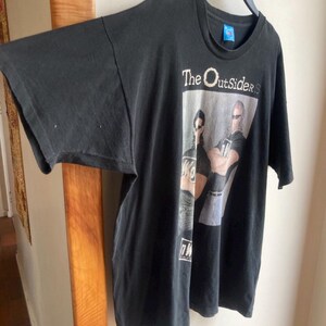 May include: Black t-shirt featuring the text "The Outsiders" above a graphic of two men wearing sunglasses. The shirt is hanging on a black hanger. The t-shirt has short sleeves and a crew neck.
