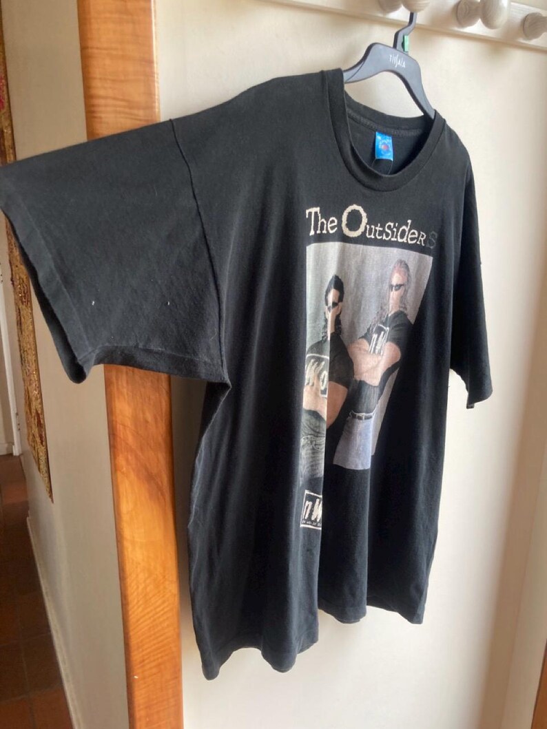 May include: Black t-shirt featuring the text "The Outsiders" above a graphic of two men wearing sunglasses. The shirt is hanging on a black hanger. The t-shirt has short sleeves and a crew neck.