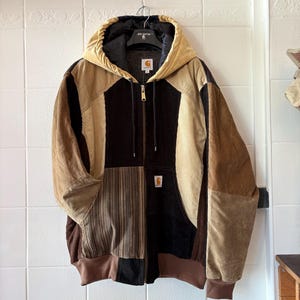 Può includere: Una giacca patchwork in velluto a coste nei toni del marrone, beige e nero è appesa a un'appendiabiti. La giacca presenta un cappuccio, una chiusura frontale con zip e una toppa con il logo Carhartt. Il design include vari pannelli in velluto a coste.