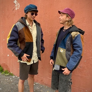 May include: Two people wearing patchwork jackets with a variety of colors including blue, brown, yellow, and gray. One person wears a blue cap and sunglasses, the other a maroon cap. Both wear shorts and sneakers or sandals. The jackets have a hooded design.