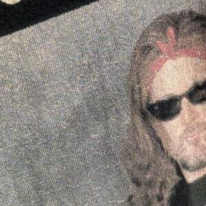May include: Close-up of a black t-shirt with a faded image of a person wearing sunglasses and a red bandana. The top of the shirt has white text. The shirt has a textured, slightly worn appearance.
