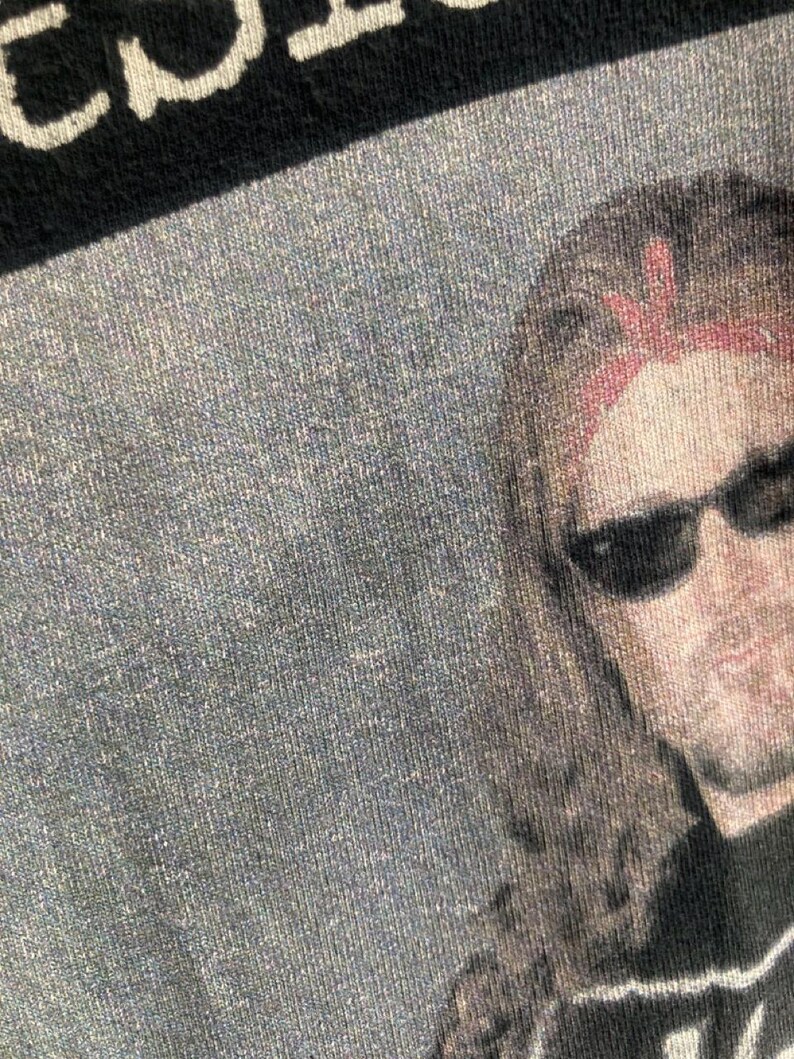 May include: Close-up of a black t-shirt with a faded image of a person wearing sunglasses and a red bandana. The top of the shirt has white text. The shirt has a textured, slightly worn appearance.