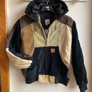 Peut inclure: Veste en velours côtelé multicolore avec capuche. Elle présente des panneaux marron, beige, noir et vert olive. Fermeture éclair et petit écusson logo. La veste est suspendue à un mur blanc. Vêtement d'extérieur.