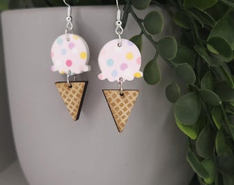 Puede incluir: Un par de pendientes colgantes con un diseño de bola de helado rosa claro con lunares coloridos, combinados con un cono de gofre de madera. Los pendientes cuelgan de ganchos plateados. Los pendientes se exhiben frente a una maceta gris con follaje verde.