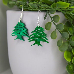 May include: A pair of green earrings shaped like cedar trees, a symbol of Lebanon. The earrings are made of a green material with a marbled effect and have silver hooks.