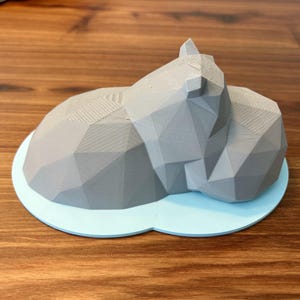 Puede incluir: Una escultura de un gato durmiendo, impresa en 3D, de color gris y geométrica, sobre una base azul claro.