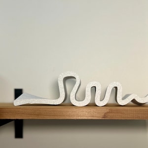 May include: A white, abstract sculpture with a wavy design. The textured piece is displayed on a wooden shelf with black metal brackets, against a neutral wall. The sculpture's form resembles a stylized word or shape.